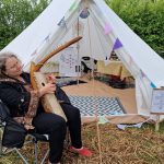 Lady sitting playing the harp outside one of the 1-2-1 healing tents at the Conscious Heart Warrior's healing village for festivals.