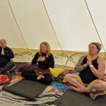 Three ladies sitting with hand to their hearts in Muladhara ceremony space at the Conscious Heart Warriors healing village for festivals.